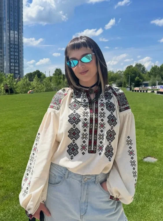 Woman in a light vyshyvanka with a red and black ornament and denim shorts on a city lawn