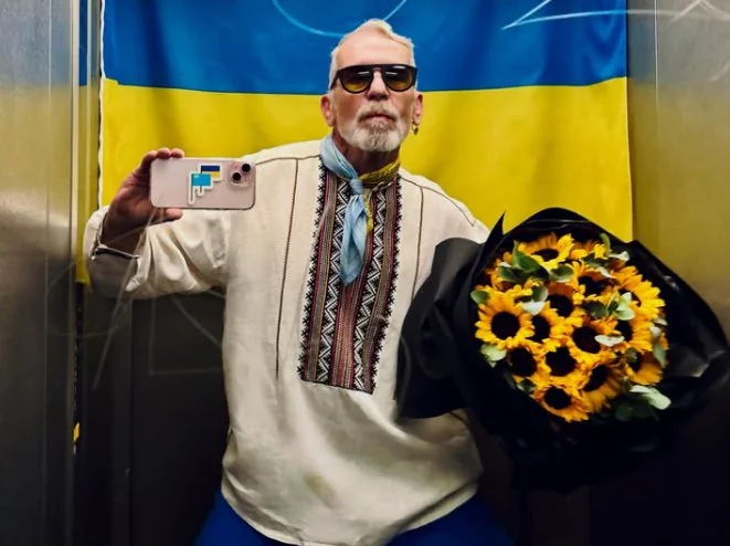 Man in a vyshyvanka with a bouquet of sunflowers taking a selfie against the Ukrainian flag